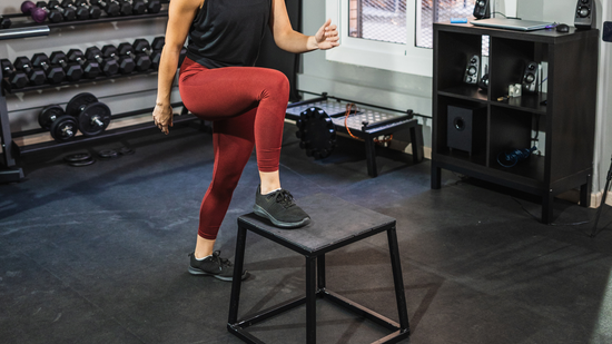 Person stepping onto a black plyo box in a gym, wearing red leggings and black top.