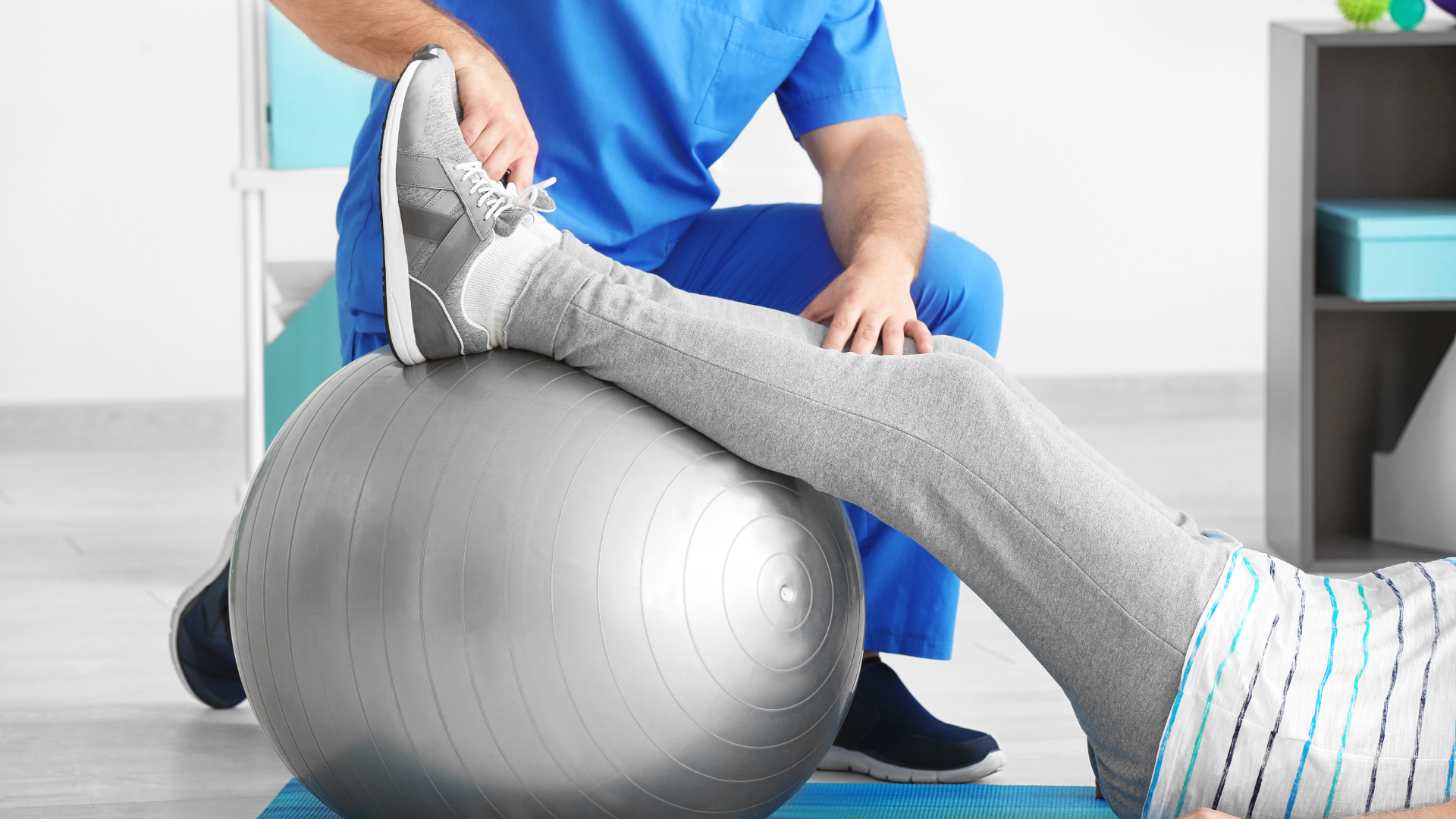 Physical therapist stretching a patient’s leg over a silver exercise ball in a clinic.