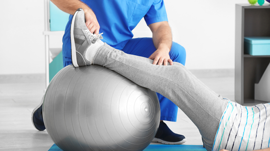Physical therapist stretching a patient’s leg over a silver exercise ball in a clinic.