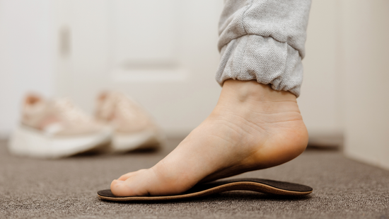 Bare foot pressing down on a collapsing brown sandal indoors, with another pair blurred in the background