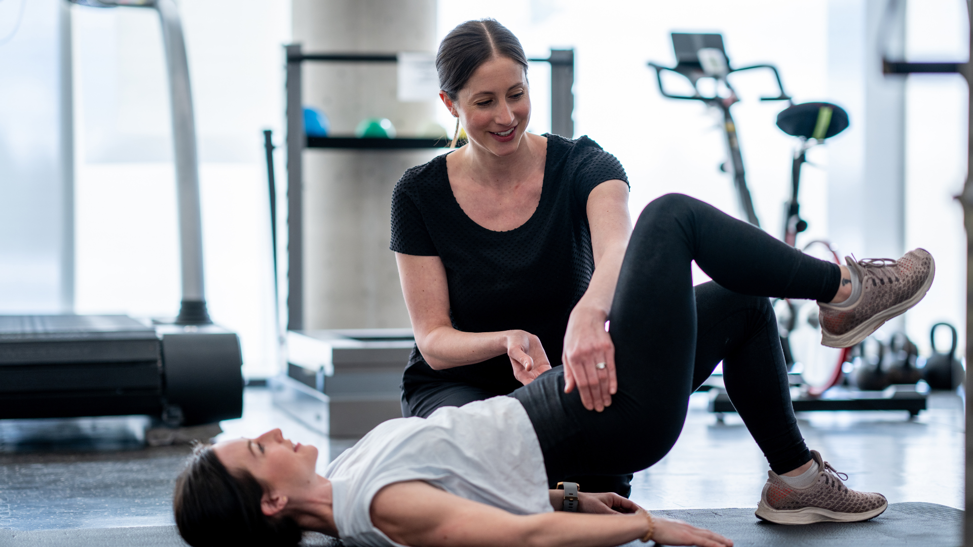 Trainer stretching a client’s leg on a mat in a bright gym.