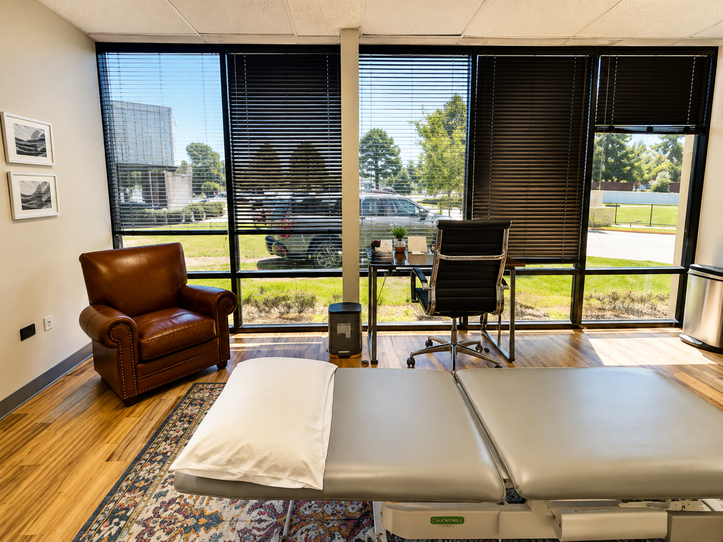 Massage room with table, leather chair, desk, and large windows overlooking a parking lot