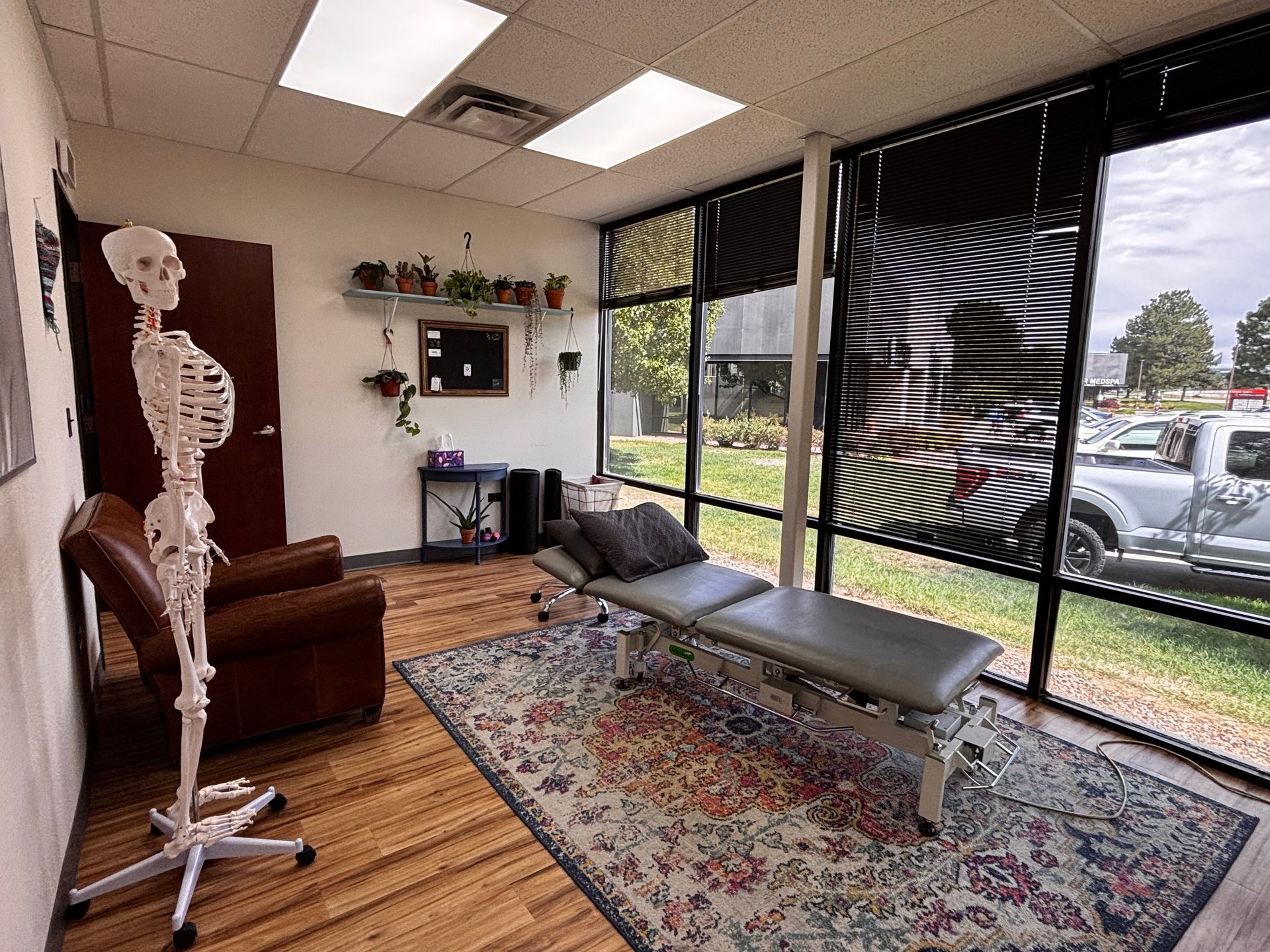 Massage room with table, leather chair, desk, and large windows overlooking a parking lot