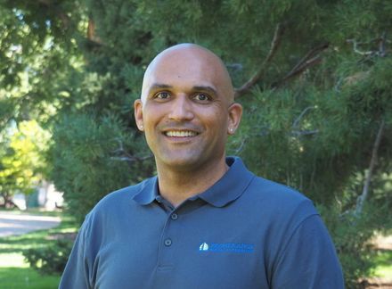 Smiling bald man in a blue polo shirt standing outdoors in front of green trees