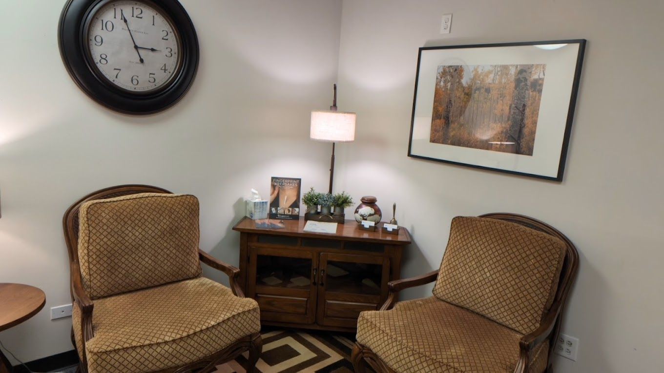Small waiting area with two wicker chairs, a wood side table, lamp, wall clock, and framed artwork.