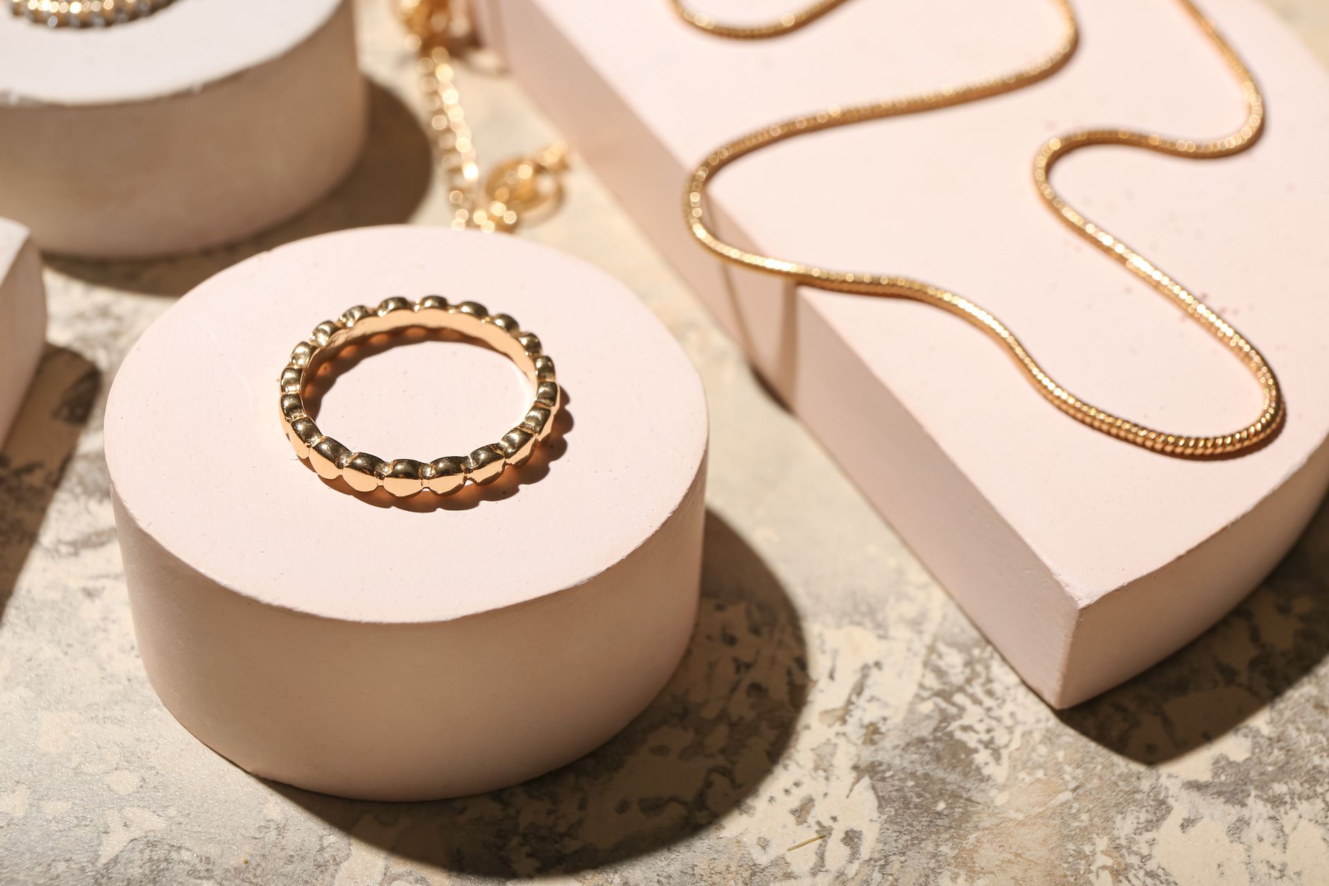 Gold jewelry on pastel pink display stands, including a ring and necklace.