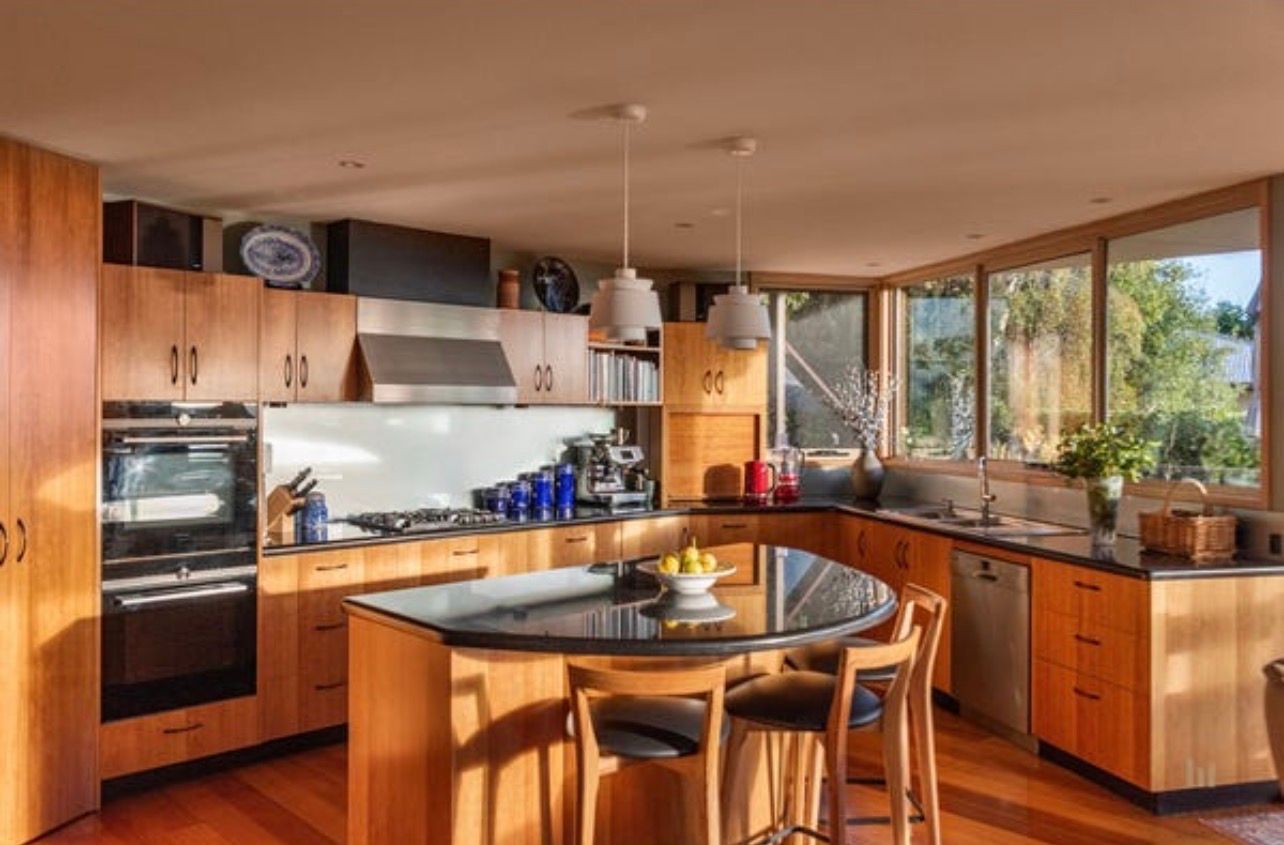 Bright modern kitchen with wooden cabinets, island table, and large windows overlooking trees.