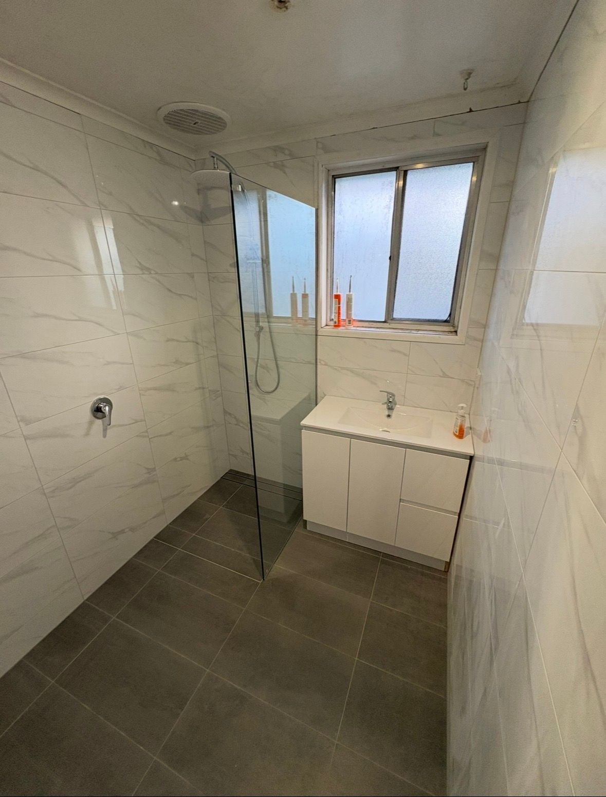Modern bathroom with glass shower, gray tile floor, white marble walls, and a window over the sink.