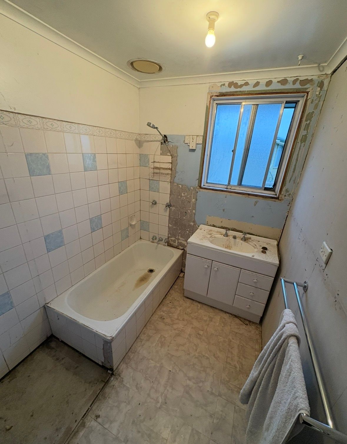 Small, worn bathroom with tub, sink cabinet, tiled walls, towel rack, and a window above the sink.