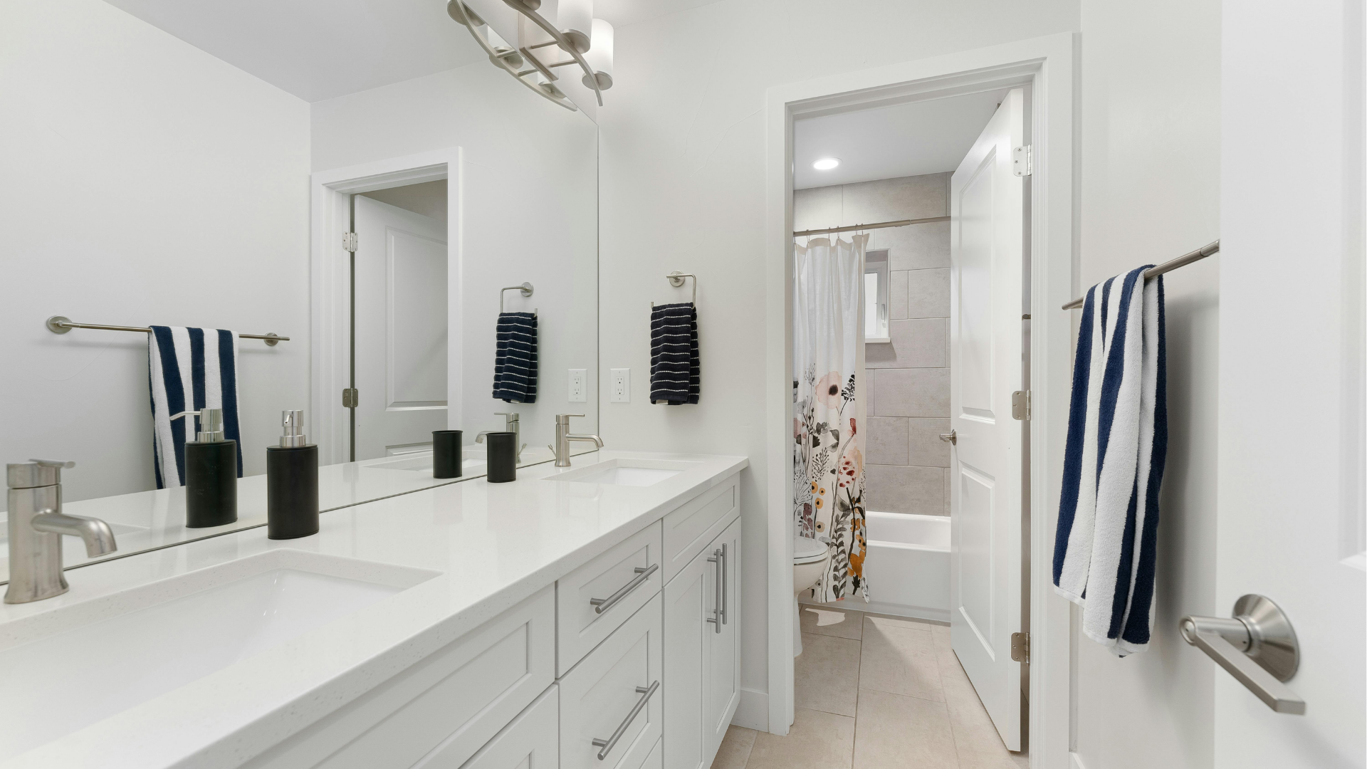 Bright white bathroom with double vanity, mirrors, towels, and an open doorway to a tiled shower area.