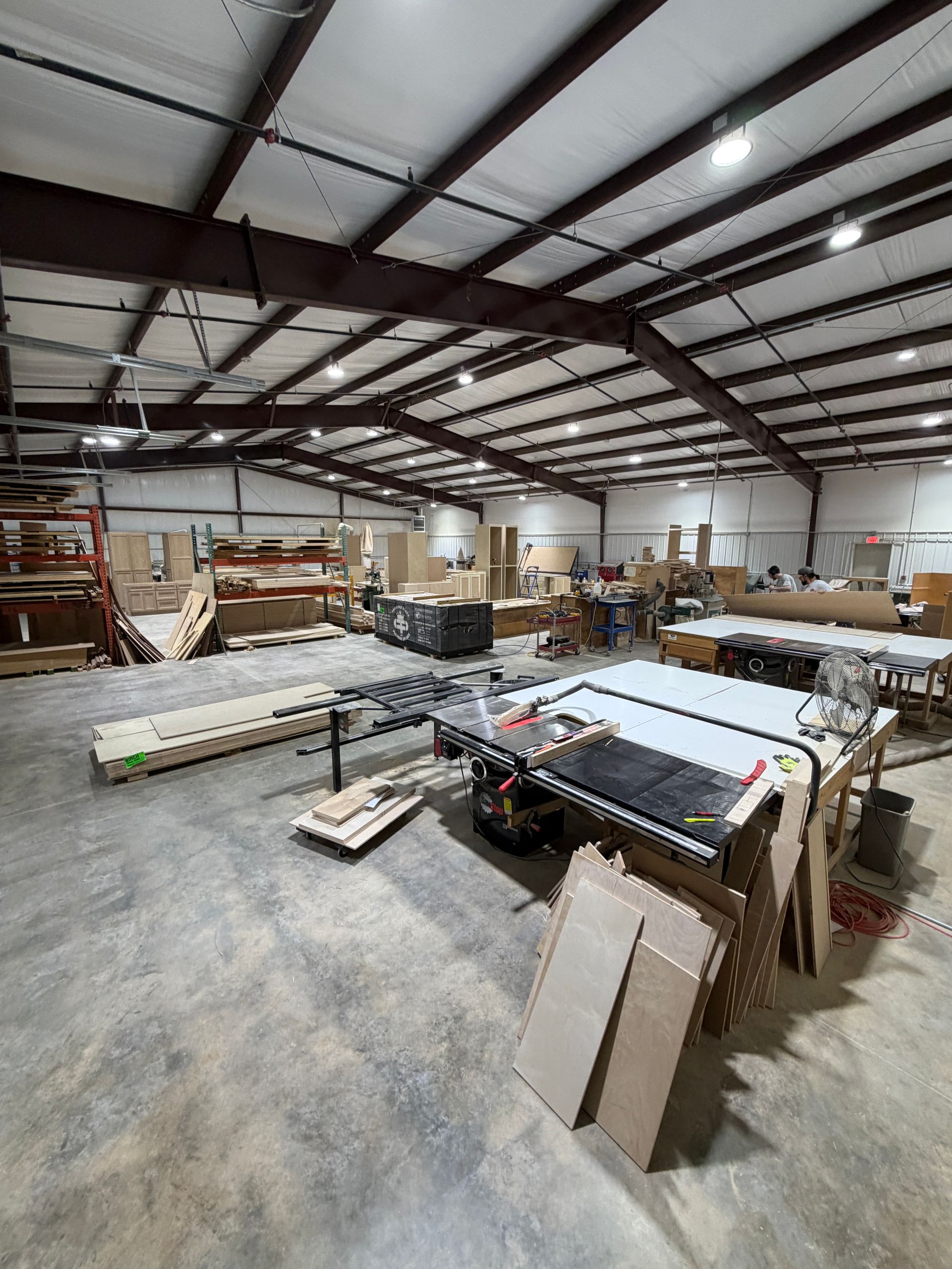 Large warehouse workshop with stacks of lumber, worktables, and tools under a metal ceiling