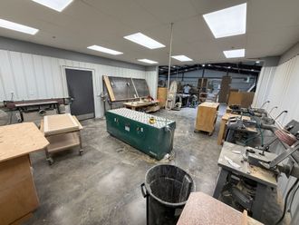 Cluttered workshop with tools, workbenches, cardboard boxes, and a trash can under bright ceiling lights