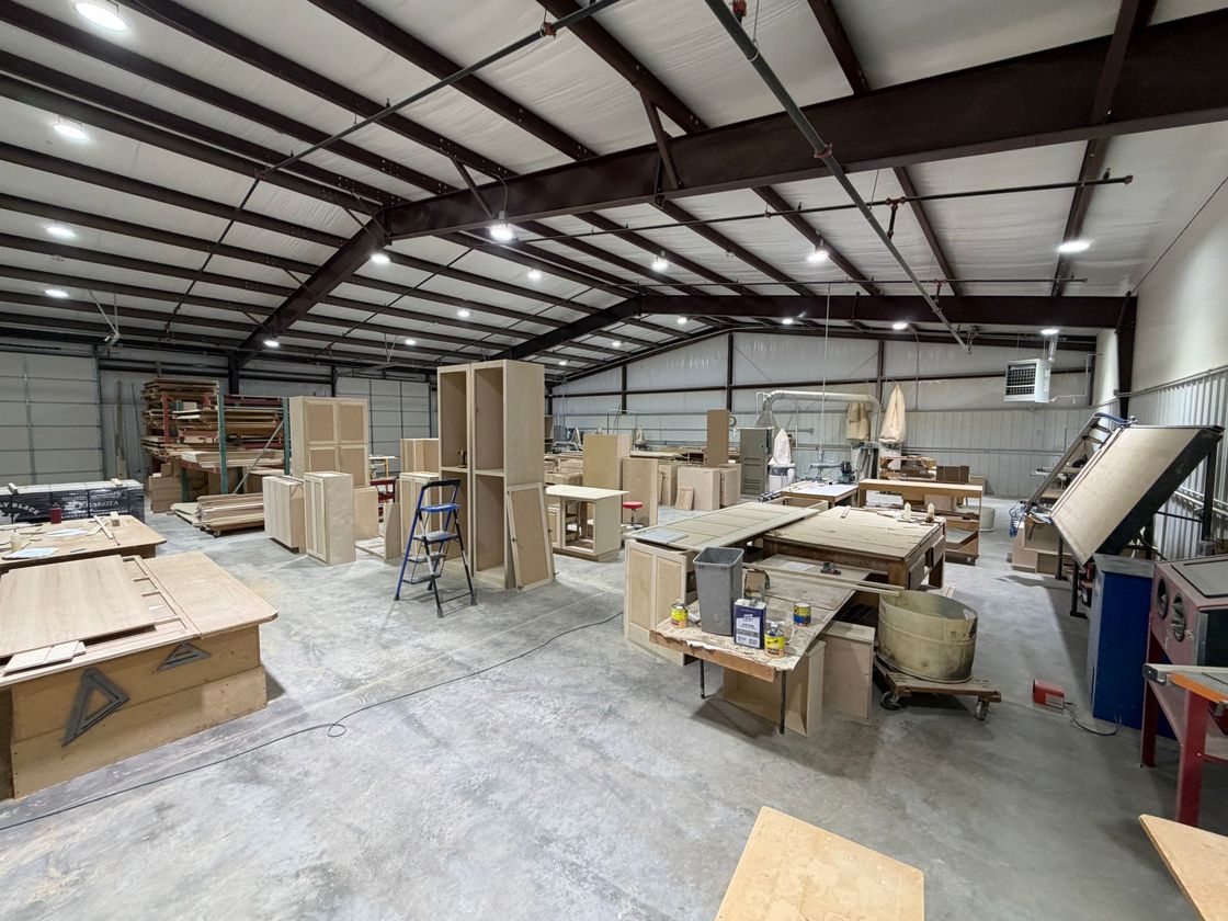 Spacious woodworking shop with tables, cabinets, tools, and lumber under a high metal ceiling