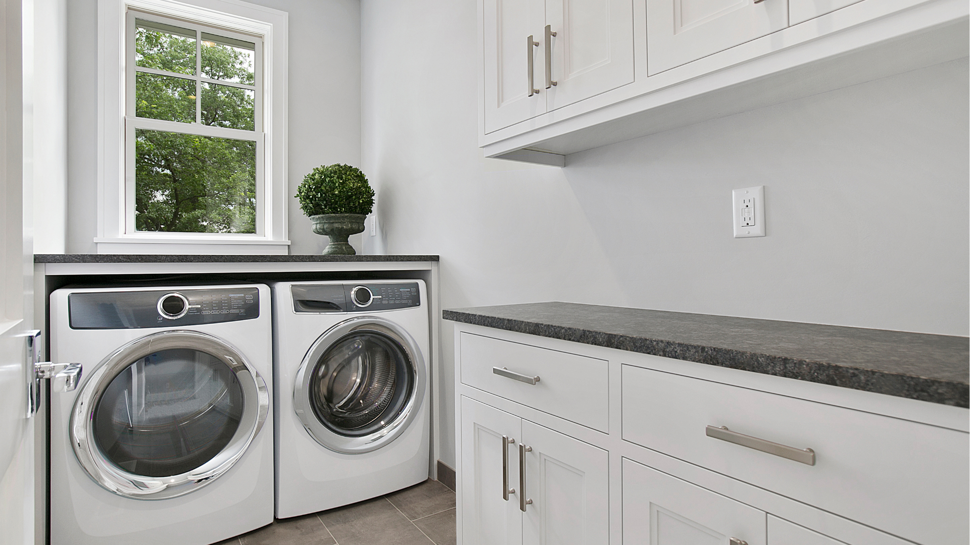 Bright laundry room with washer, dryer, white cabinets, and a window with a plant.