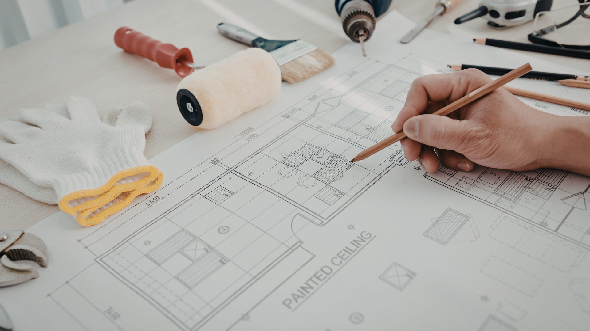 Hands sketching a home design blueprint with pencil, ruler, paint roller, and tools on a desk