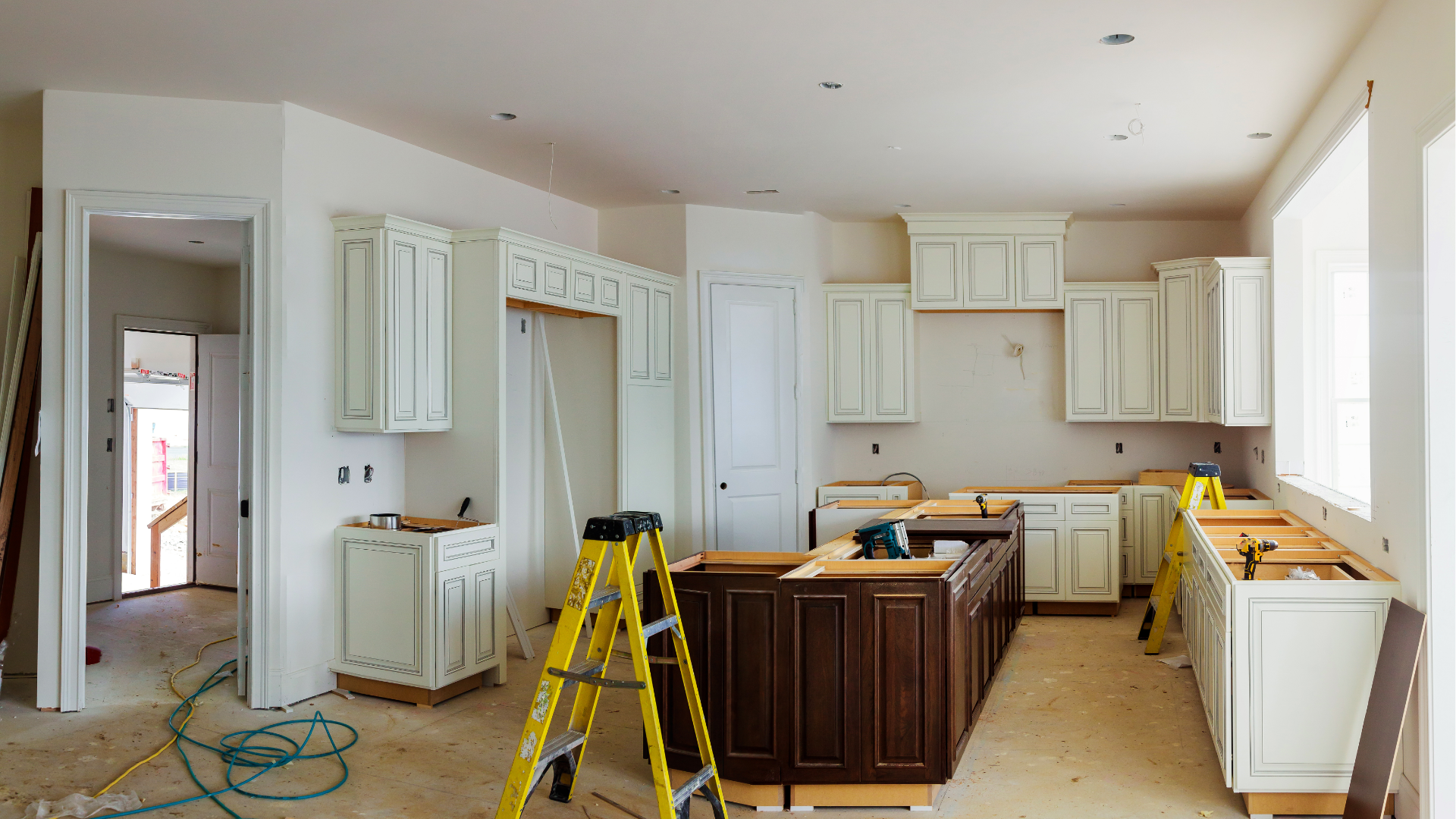 Unfinished kitchen renovation with white cabinets, island, yellow ladder, and bare floors.