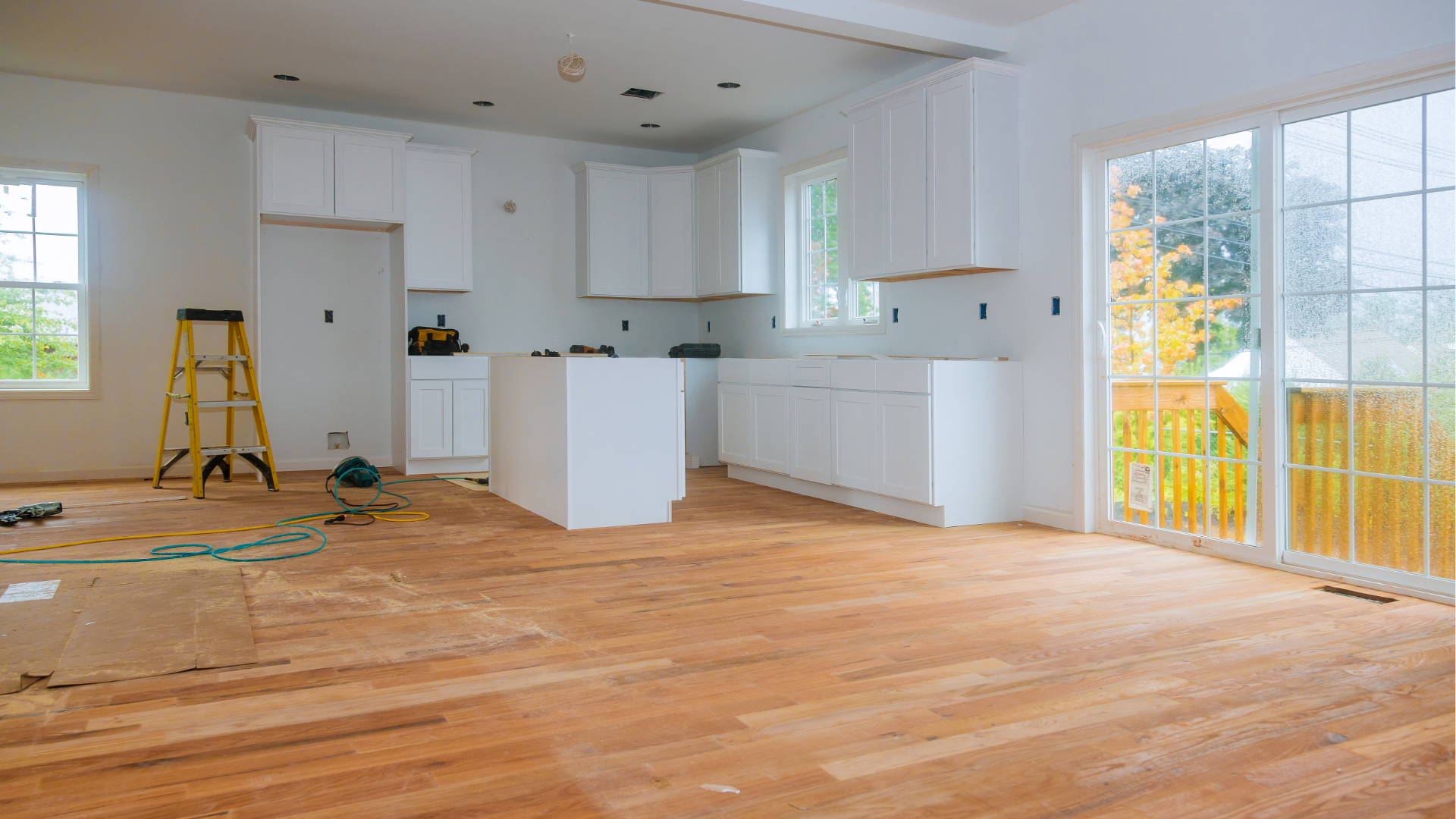Empty kitchen under renovation with white cabinets, hardwood floors, and a ladder near a sliding glass door.