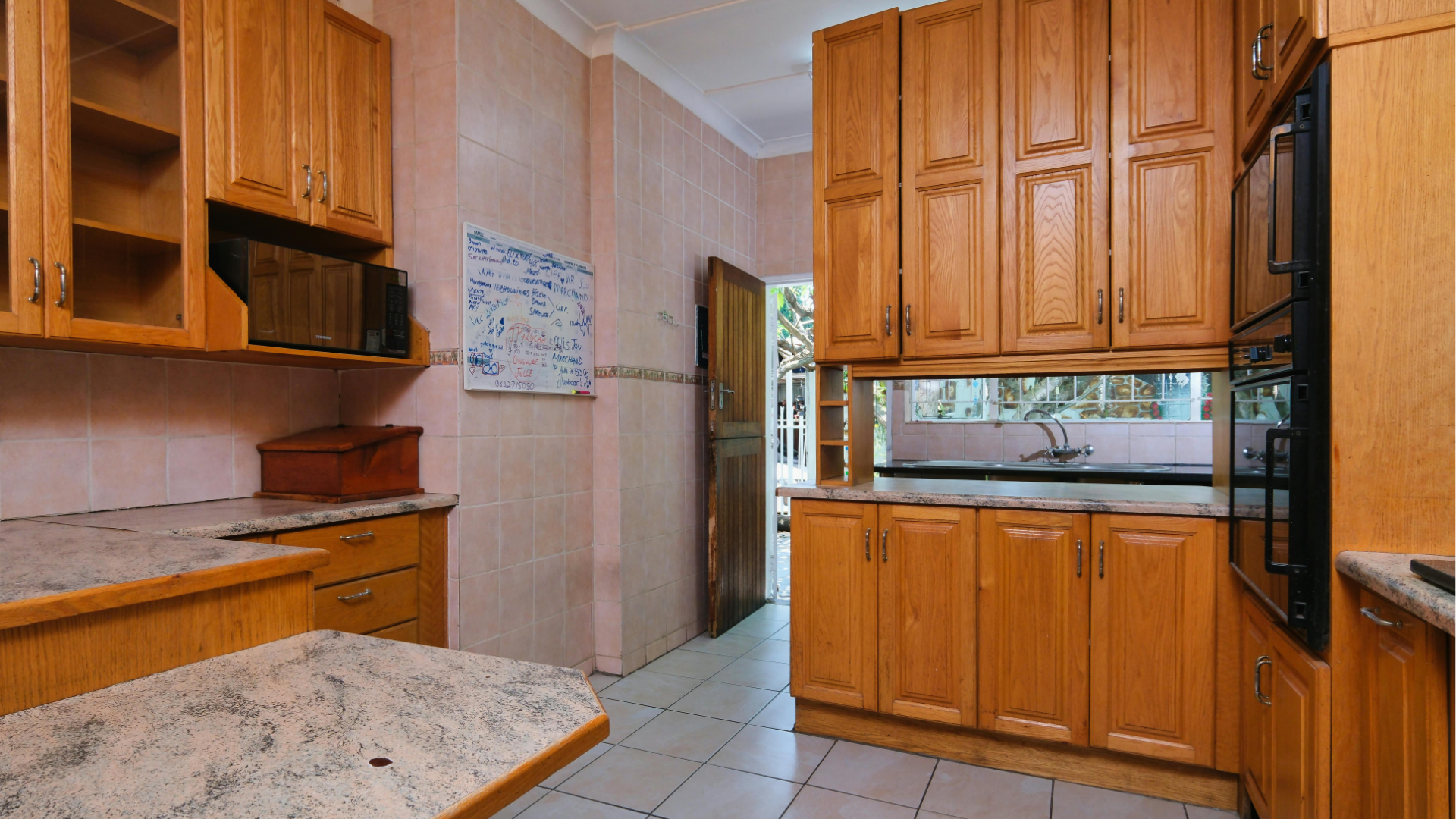 Empty kitchen with wood cabinets, granite counters, and white tile floor, viewed toward a doorway.