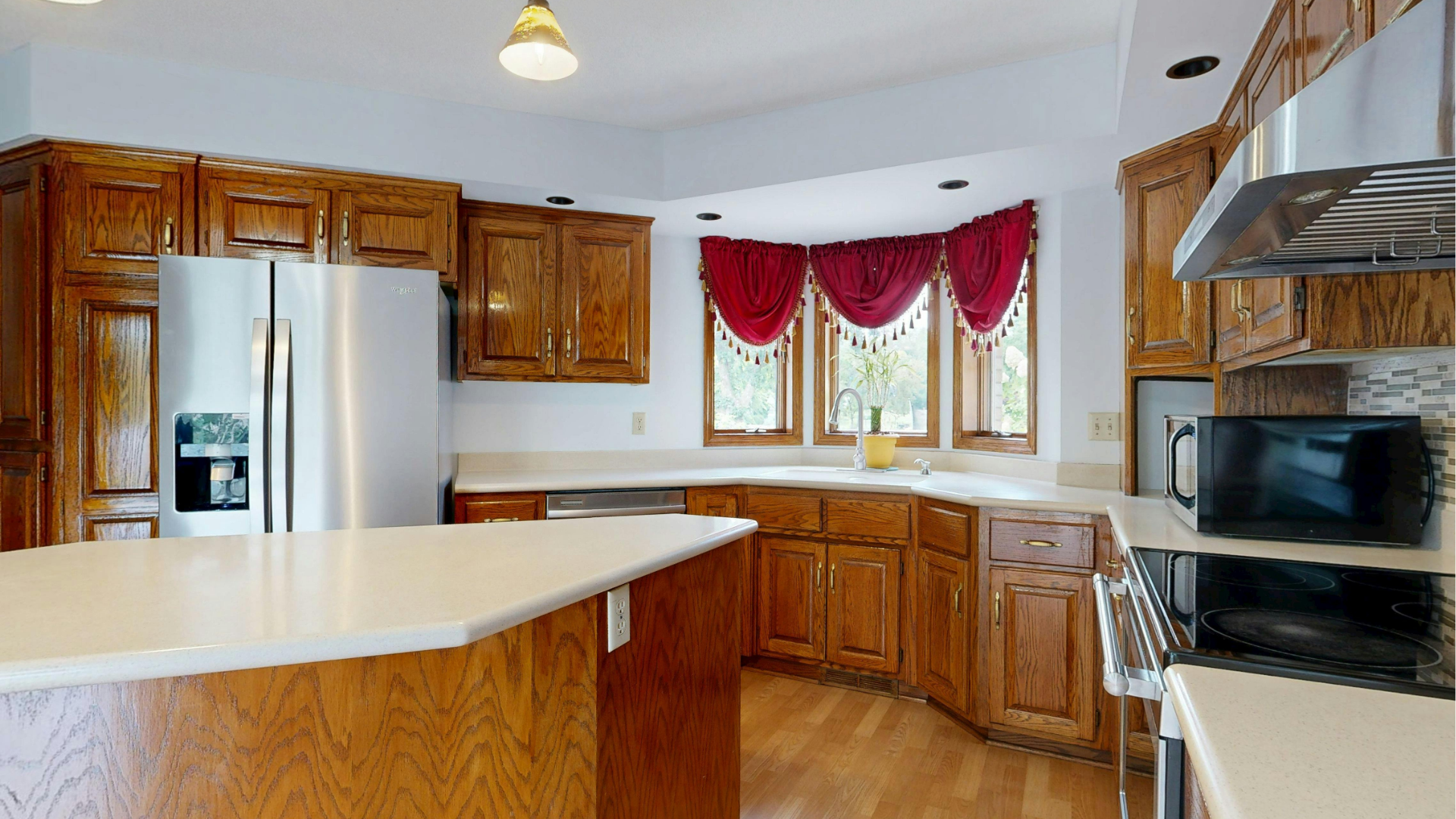 Bright kitchen with wooden cabinets, stainless fridge, white countertops, and red window valances.