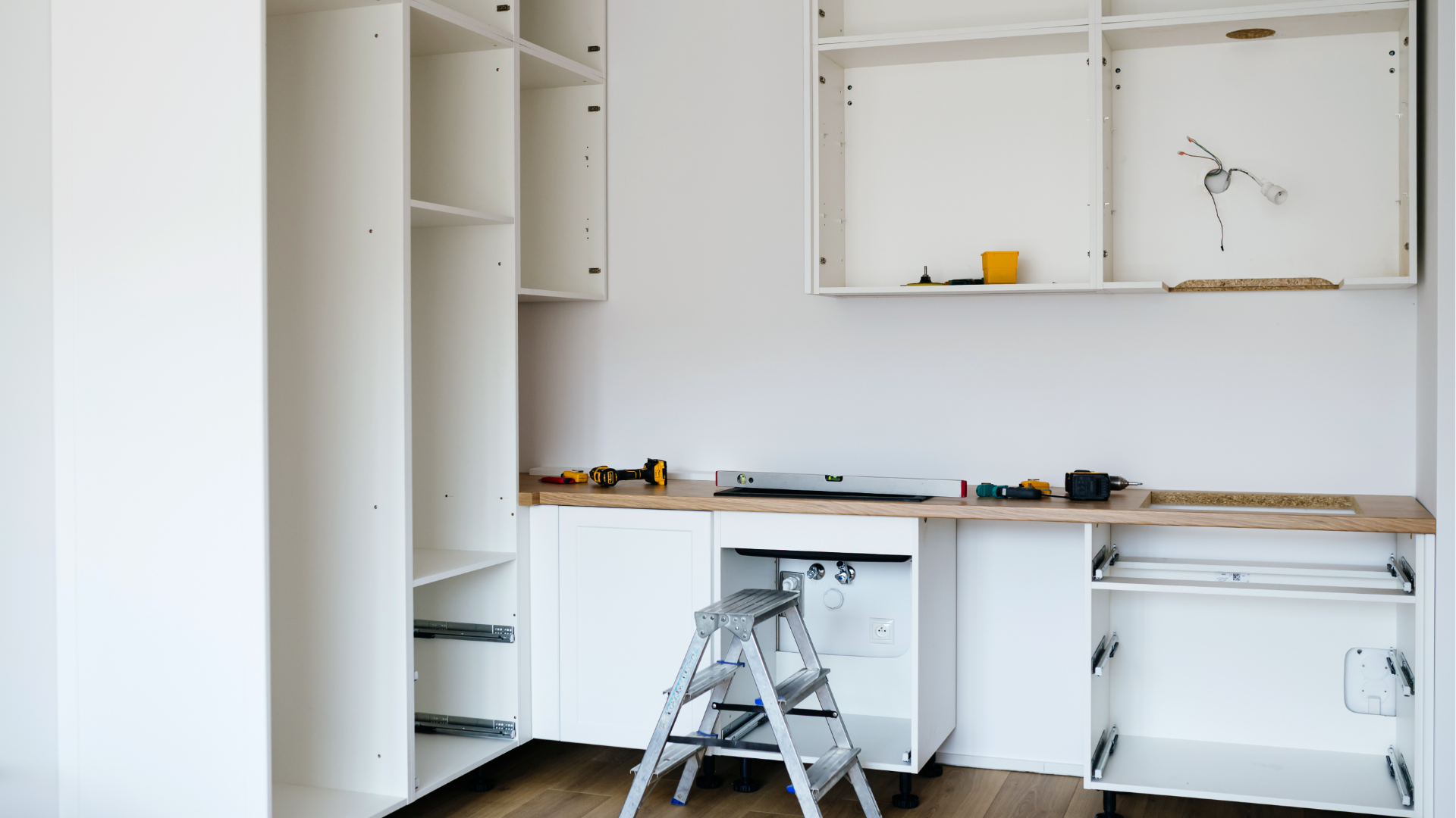 Partially built white office cabinets with a wooden countertop and a step ladder in a bright room