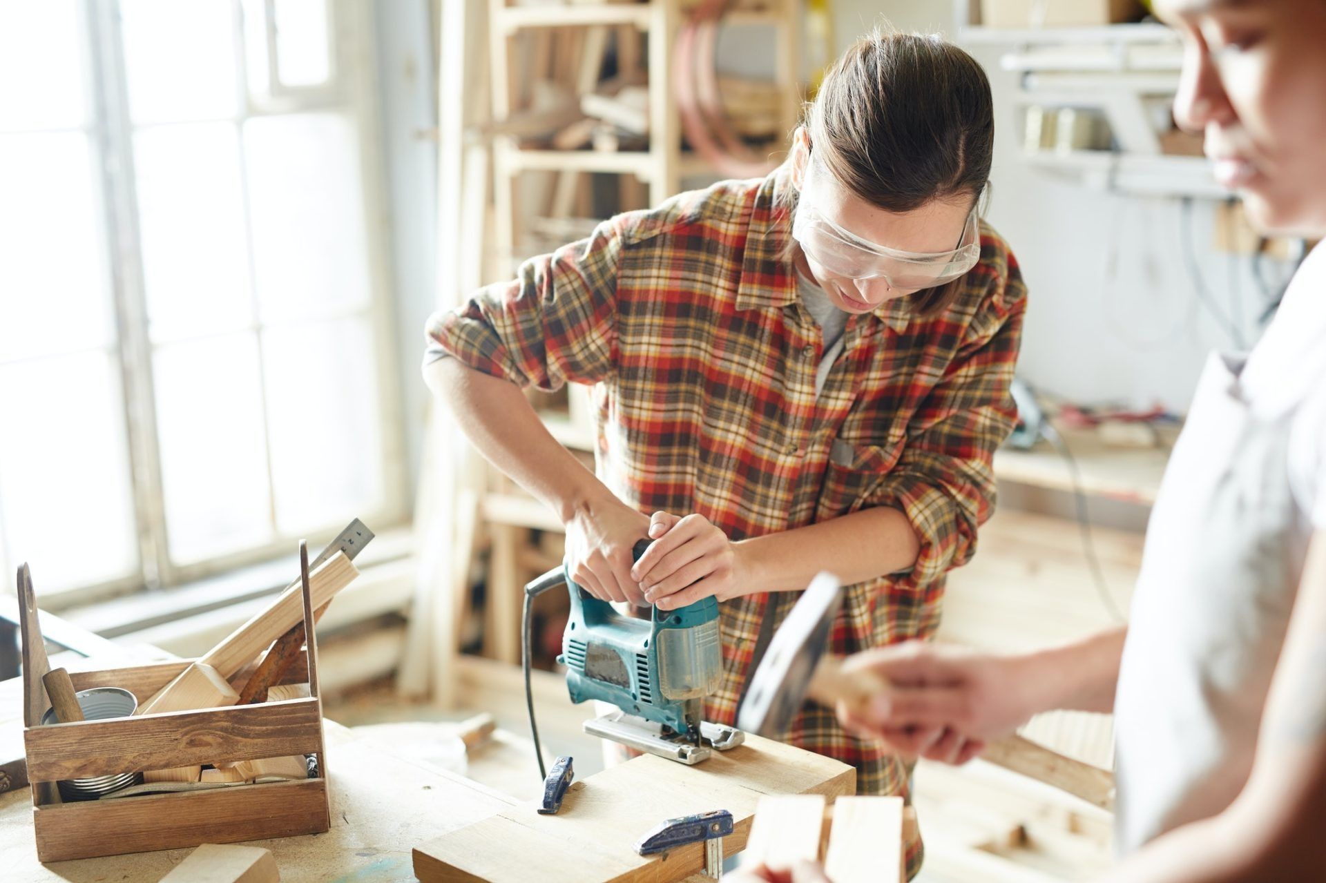 Person using a jigsaw on wood in a bright workshop, with tools and a helper nearby.