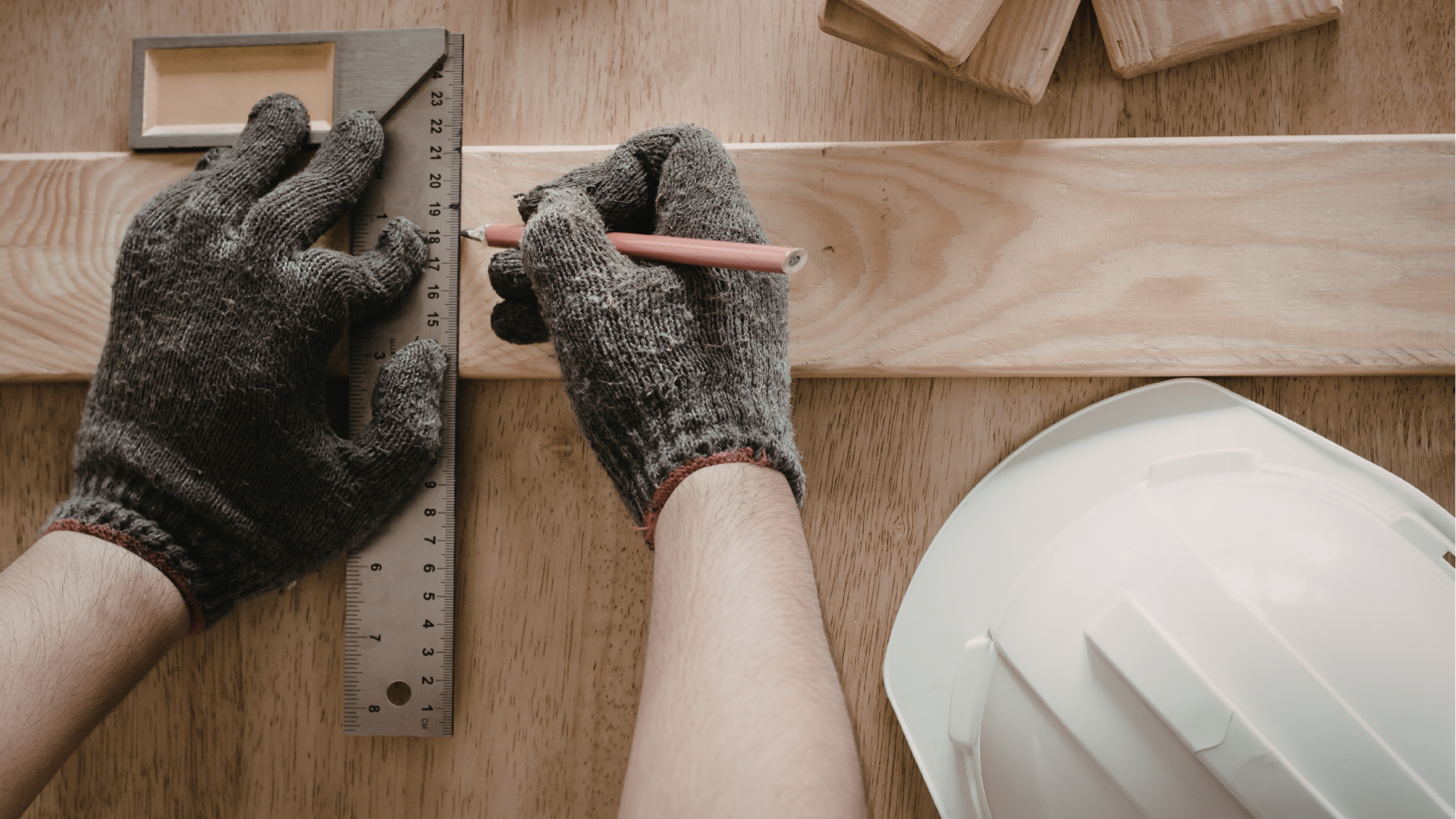 Gloved hands measuring and marking wood with a pencil, with a hard hat on a workbench.