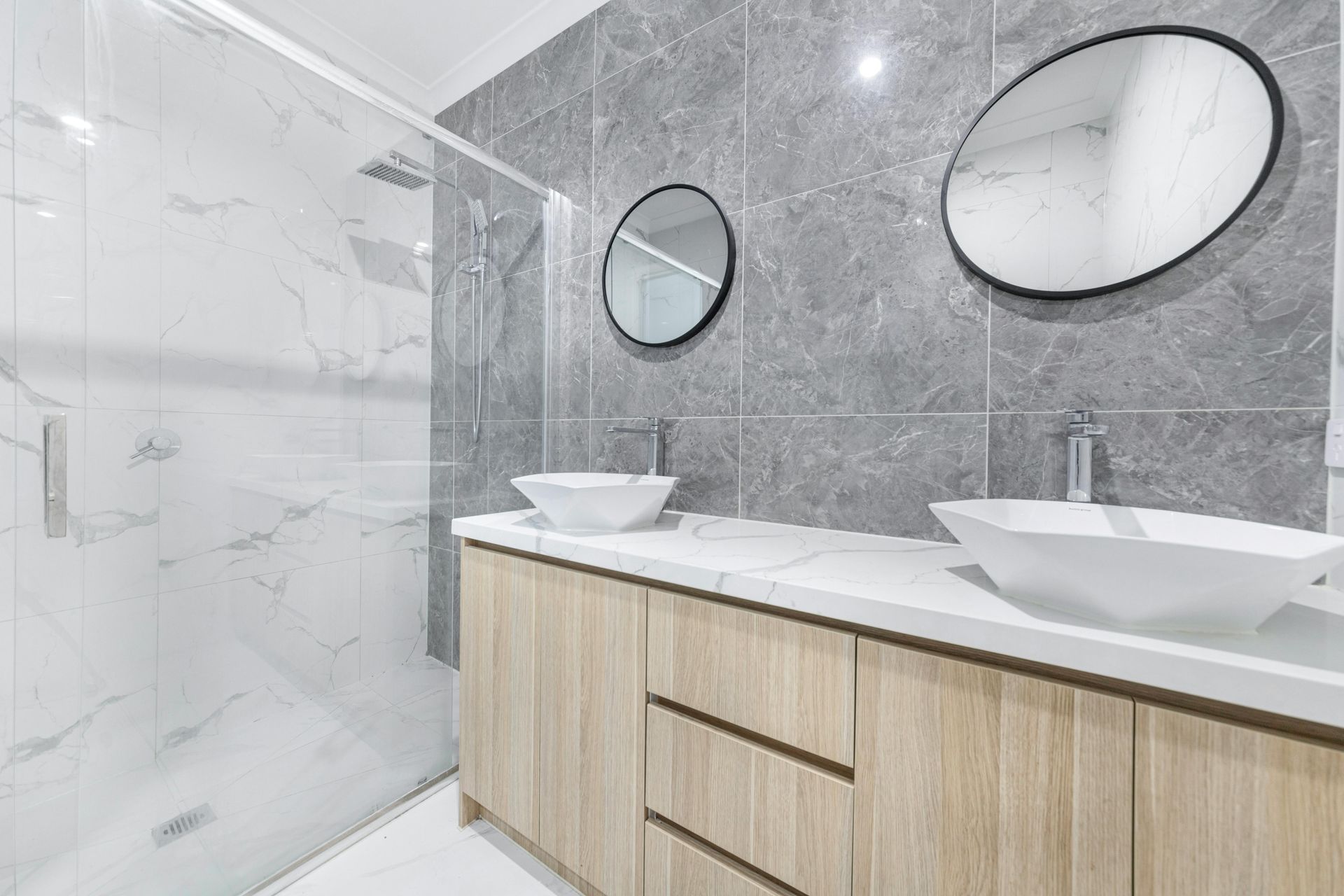 Modern bathroom with dual sinks, marble walls, wood vanity, and round mirrors