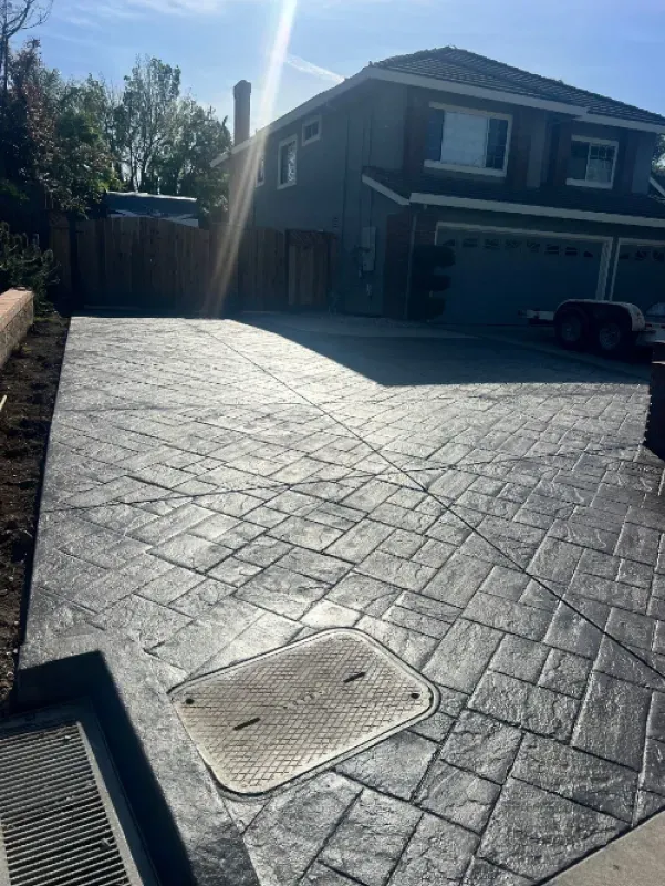 Stamped concrete driveway in front of a suburban house, with strong sunlight and a drain grate in the foreground