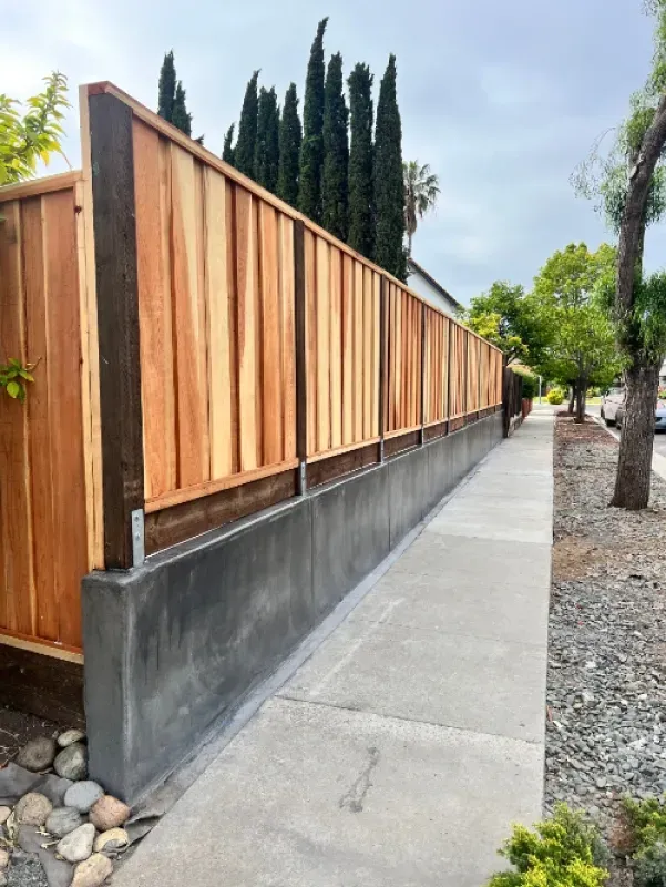 Wooden fence atop a concrete wall beside a sidewalk with trees and rocks along the edge