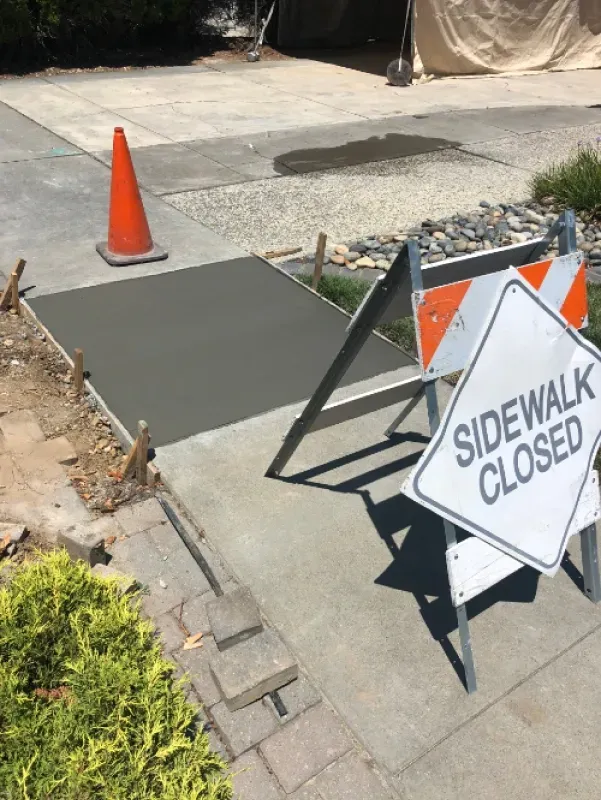 Sidewalk closed sign beside a freshly patched sidewalk and orange traffic cone