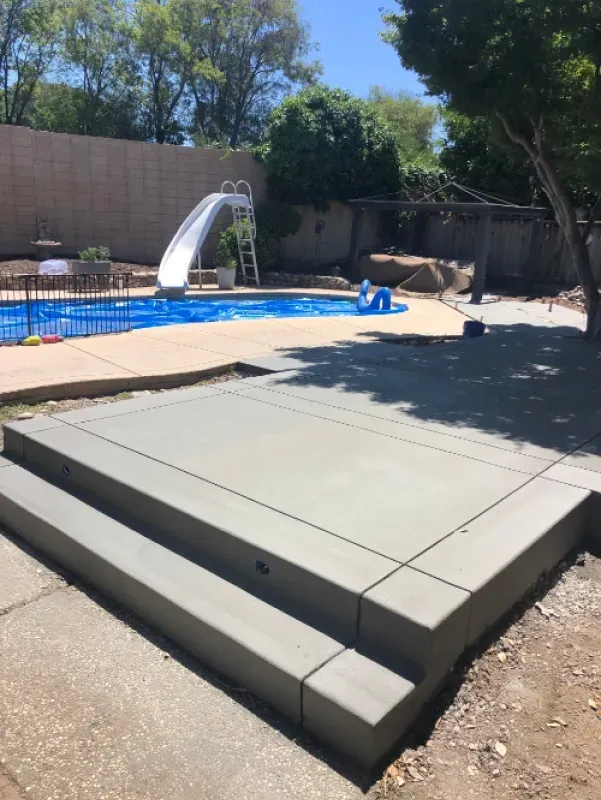 Backyard patio with raised concrete platform beside a swimming pool and shade trees