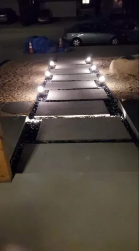 Nighttime view of a lit stone walkway leading from a driveway toward a parked car.