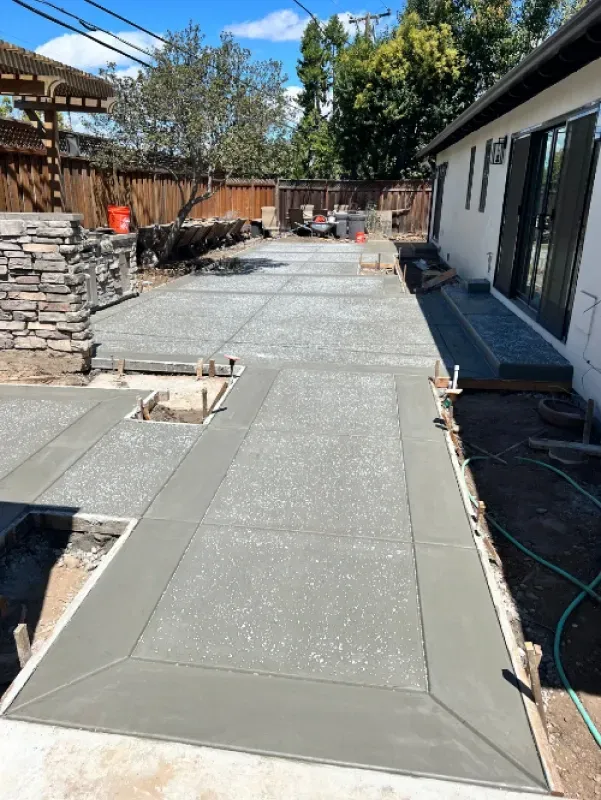 Freshly poured concrete walkway under construction beside a house and stone retaining wall, with forms and tools visible