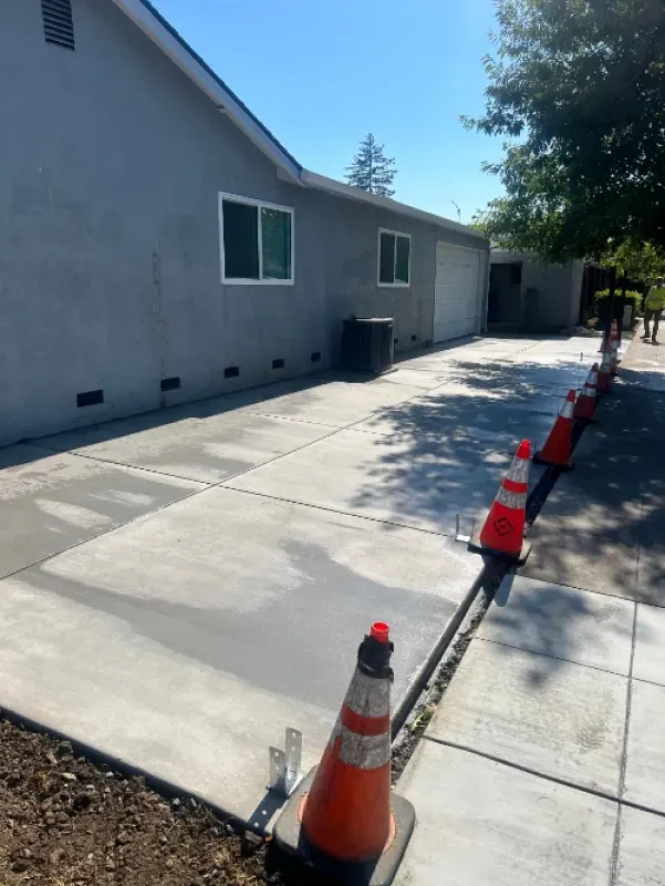 Concrete driveway beside gray building, lined with orange traffic cones in bright sunlight.