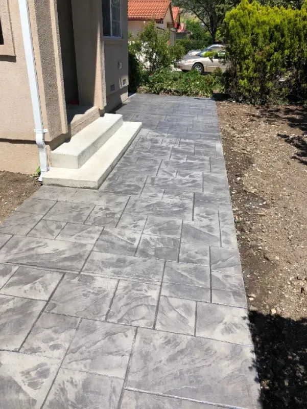 Gray tiled patio beside a house, with gravel edging and plants along the side yard.
