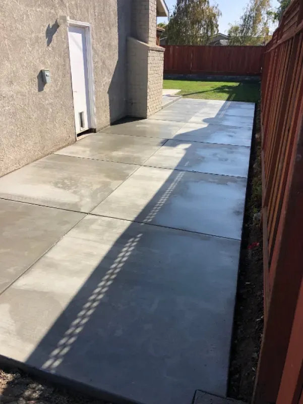 Concrete side yard walkway beside a house and red fence, with strong sunlight casting shadows.