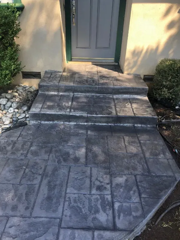 Stone walkway leading to a gray front door, bordered by rocks and shrubs, with sunlight and shadows on the path