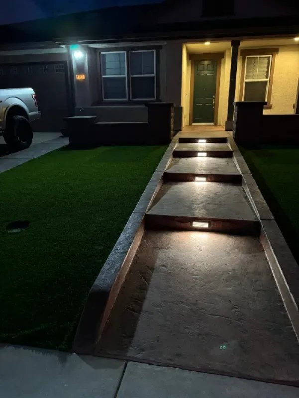 Front walkway with lit steps leading to a house entrance at night, beside a parked truck and green lawn