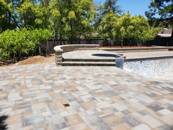 Empty backyard patio with stone pavers, curved raised seating, and a pool edge beside trees