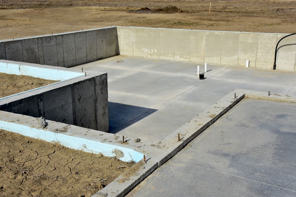 Concrete foundation slab and walls at a construction site, with exposed dirt and open rectangular cutouts.