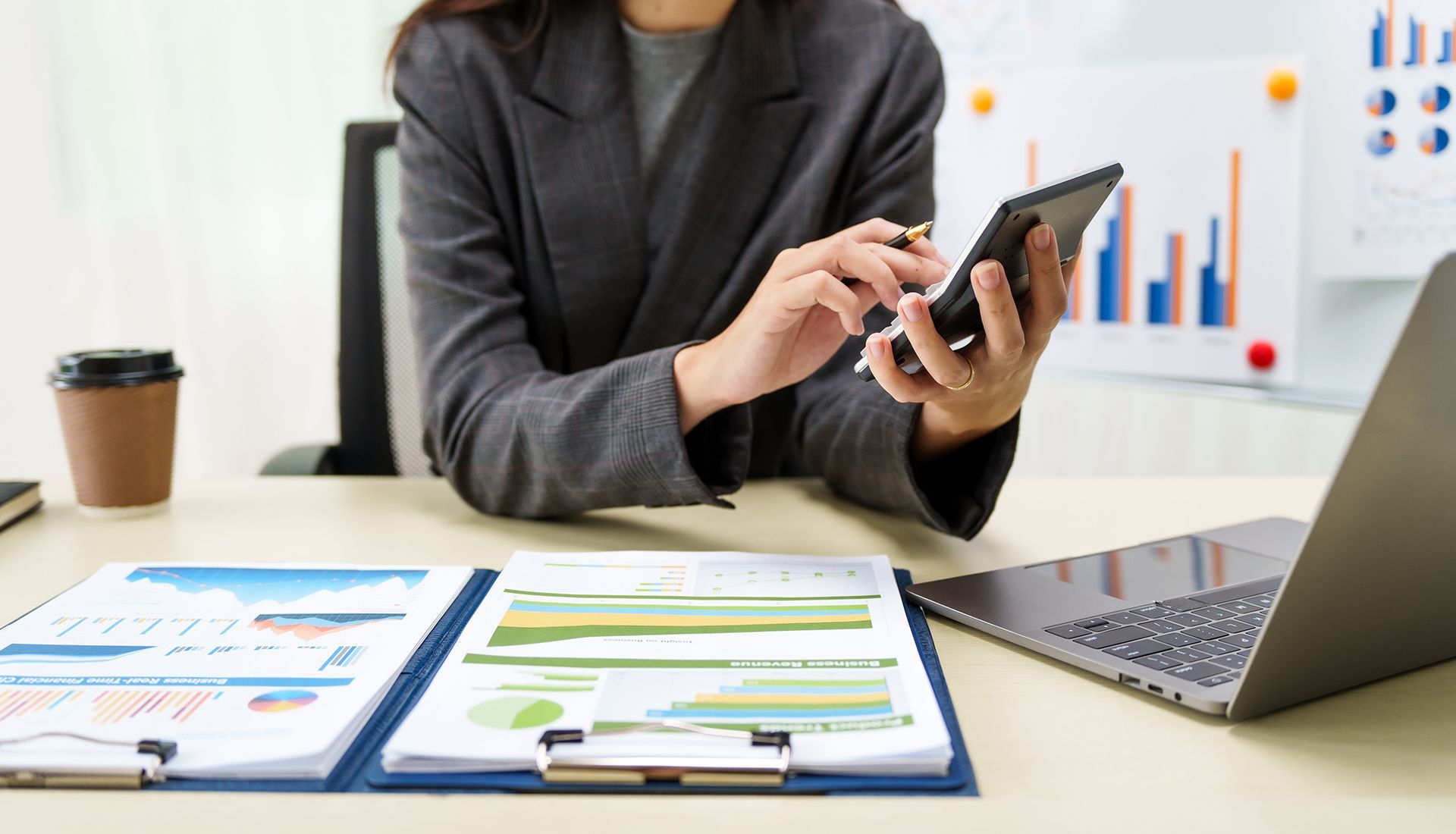 Person using a tablet at a desk with charts, documents, and an open laptop in an office