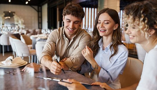 Three people smiling around a restaurant table; one writing, two observing.