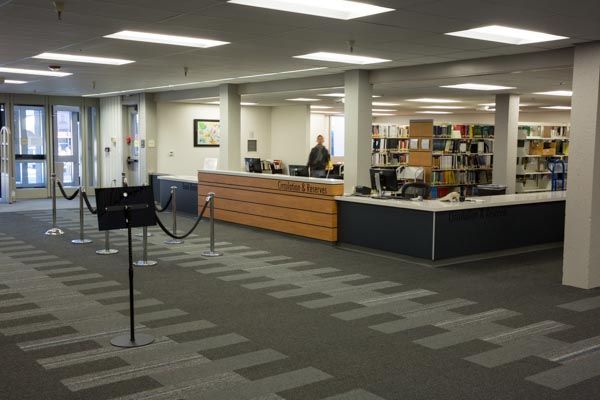 Library checkout desk with bookshelves, carpeted floor, and a person behind the counter