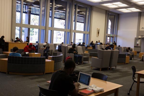 Quiet library lounge with people studying and using laptops by large windows.