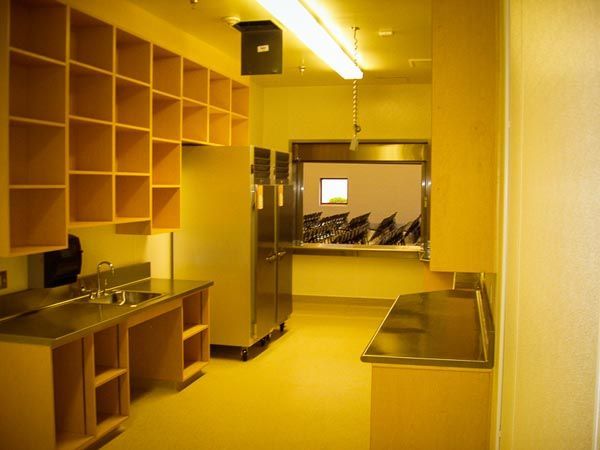 Yellow-toned small room with shelves, desks, cabinets, and a window opening to another room
