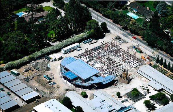 Aerial view of a construction site with a blue-roofed building, steel framework, and surrounding trees.