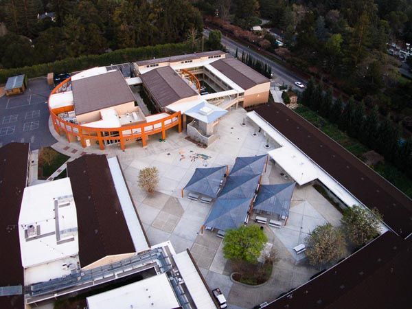 Aerial view of a modern building complex with white roofs, orange accents, and surrounding trees