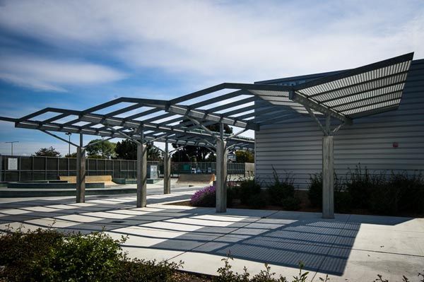 Modern outdoor patio with steel pergola, slatted shade roof, and landscaped planters in sunlight