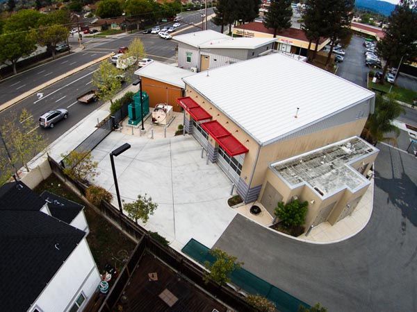 Aerial view of a small commercial building with a white roof, red awnings, and a paved parking lot beside a road.