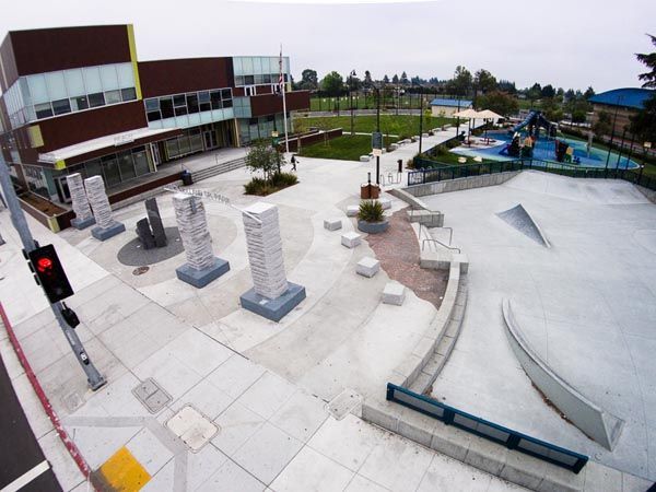 Outdoor skatepark with concrete ramps, rails, and a nearby modern building under a cloudy sky